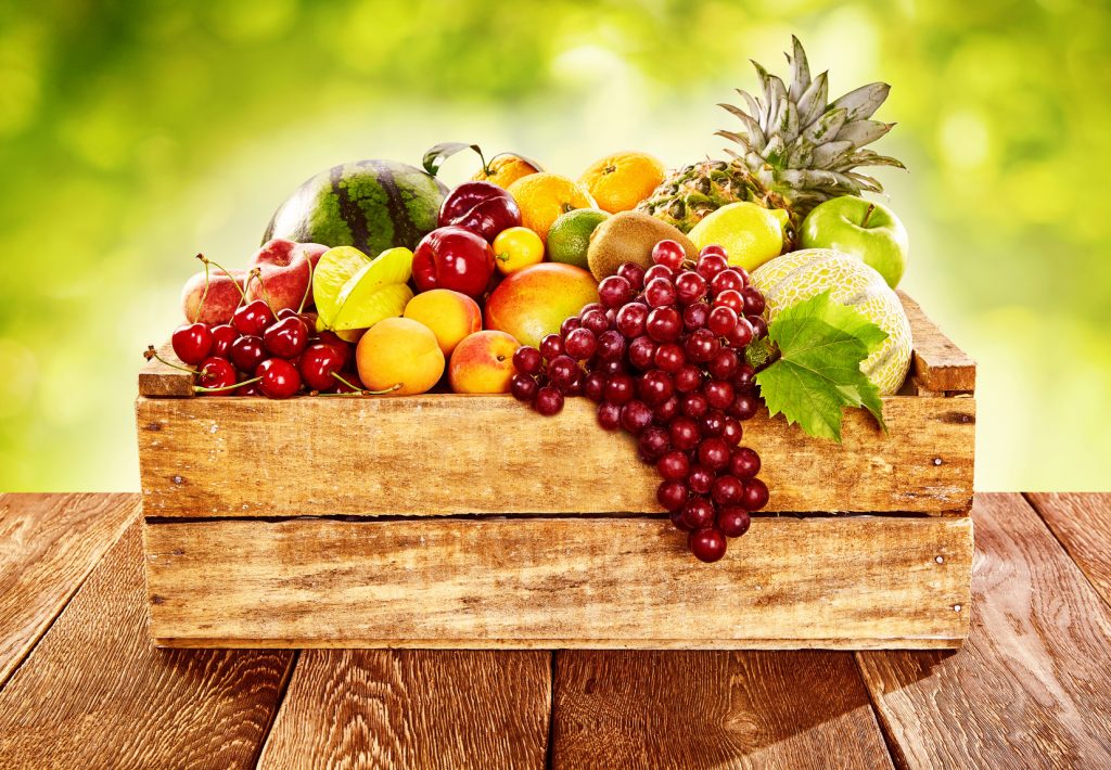 Wooden crate filled with a wide assortment of fresh healthy tropical fruit on a rustic wood table outdoors with sunlit blurred green background
