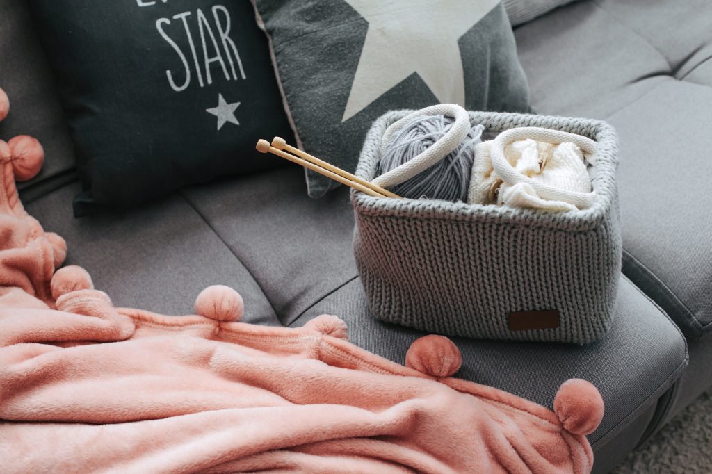 Knitted basket with yarn and needles on grey sofa by the warm pompon blanket and cushions. Still life photo of nordic interior details. Cosy place in winter decorated room.